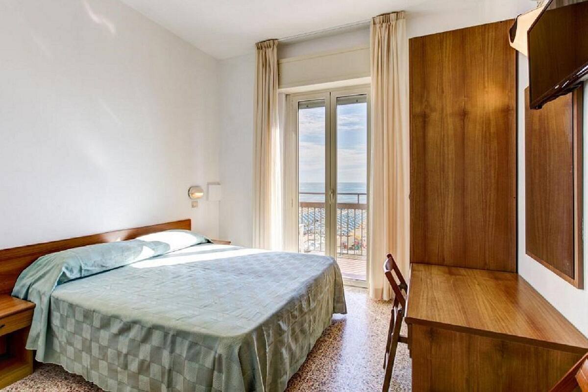 A bedroom with a bed, a desk, a chair, and a balcony with a view.
