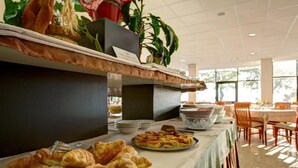 Montaje tipo bufé con croissants y otras pastelerías, un bol de fruta y una planta al fondo.