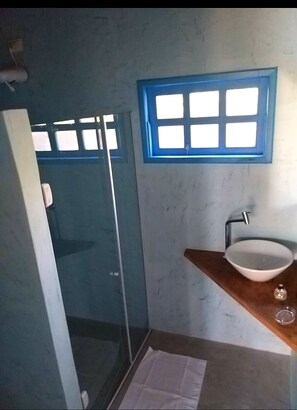 Een badkamer met een douchecabine van glas, een wit wastafeltje en een raam met blauwe rand.