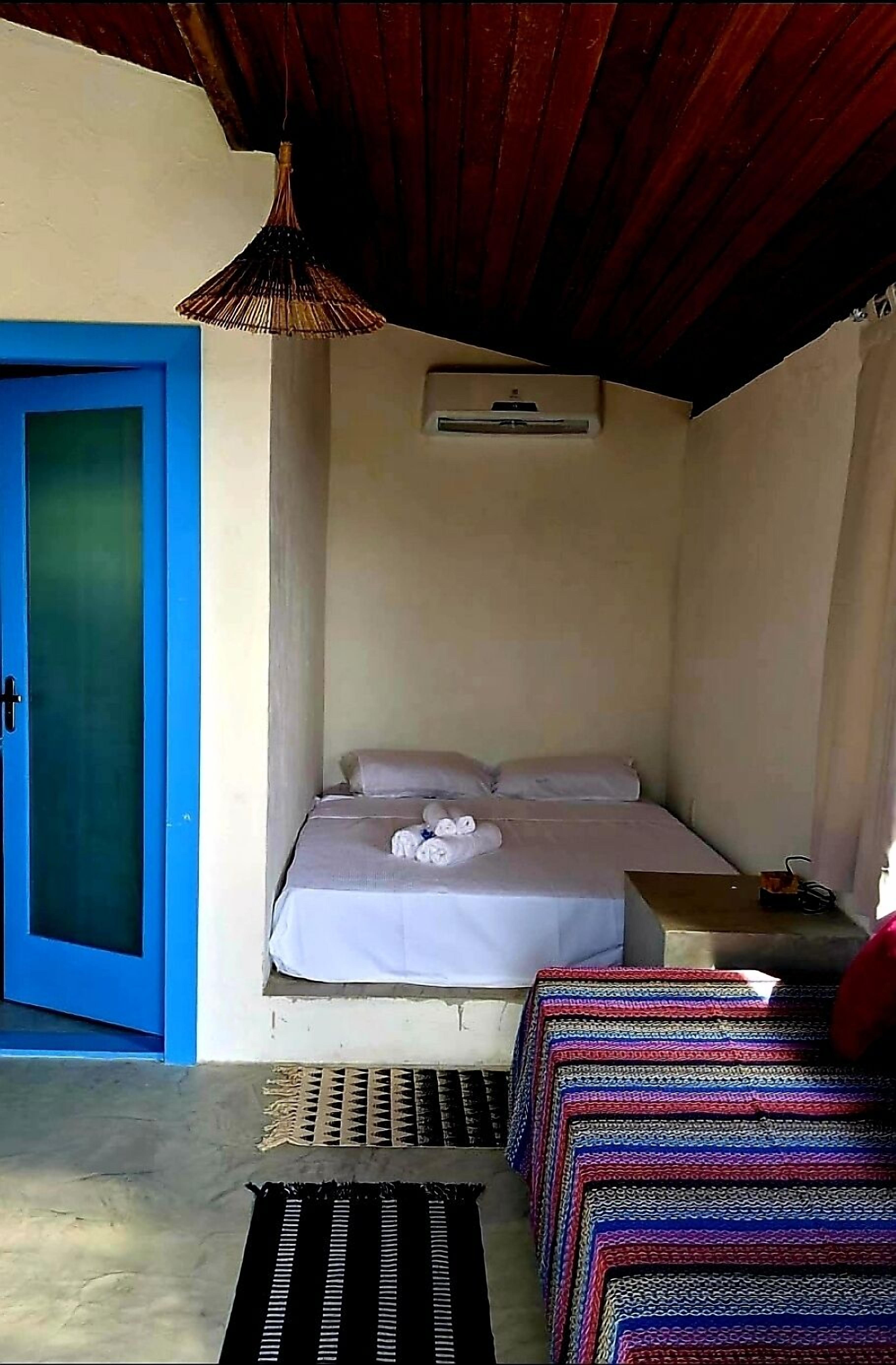 Um pequeno alojamento de um quarto, com uma cama, uma porta azul, um candeeiro pendente e uma pequena mesa com uma lâmpada.