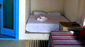 Um pequeno alojamento de um quarto, com uma cama, uma porta azul, um candeeiro pendente e uma pequena mesa com uma lâmpada.