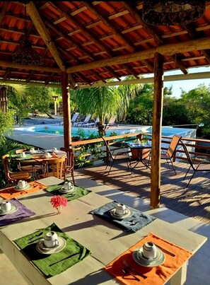 Un área de comedor al aire libre con mesas dispuestas para una comida, con vista a una piscina y abundante vegetación.