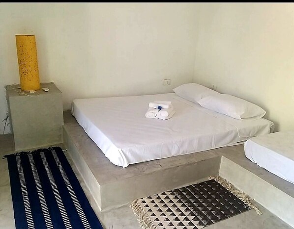 Una cama individual con ropa de cama blanca, una mesita de noche con lámpara y una alfombra estampada en el suelo.