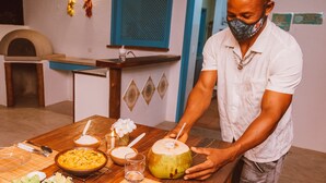 A person pouring a drink from a coconut into a glass. There is a bowl of food, a plate with a banana, and a bottle on the table.