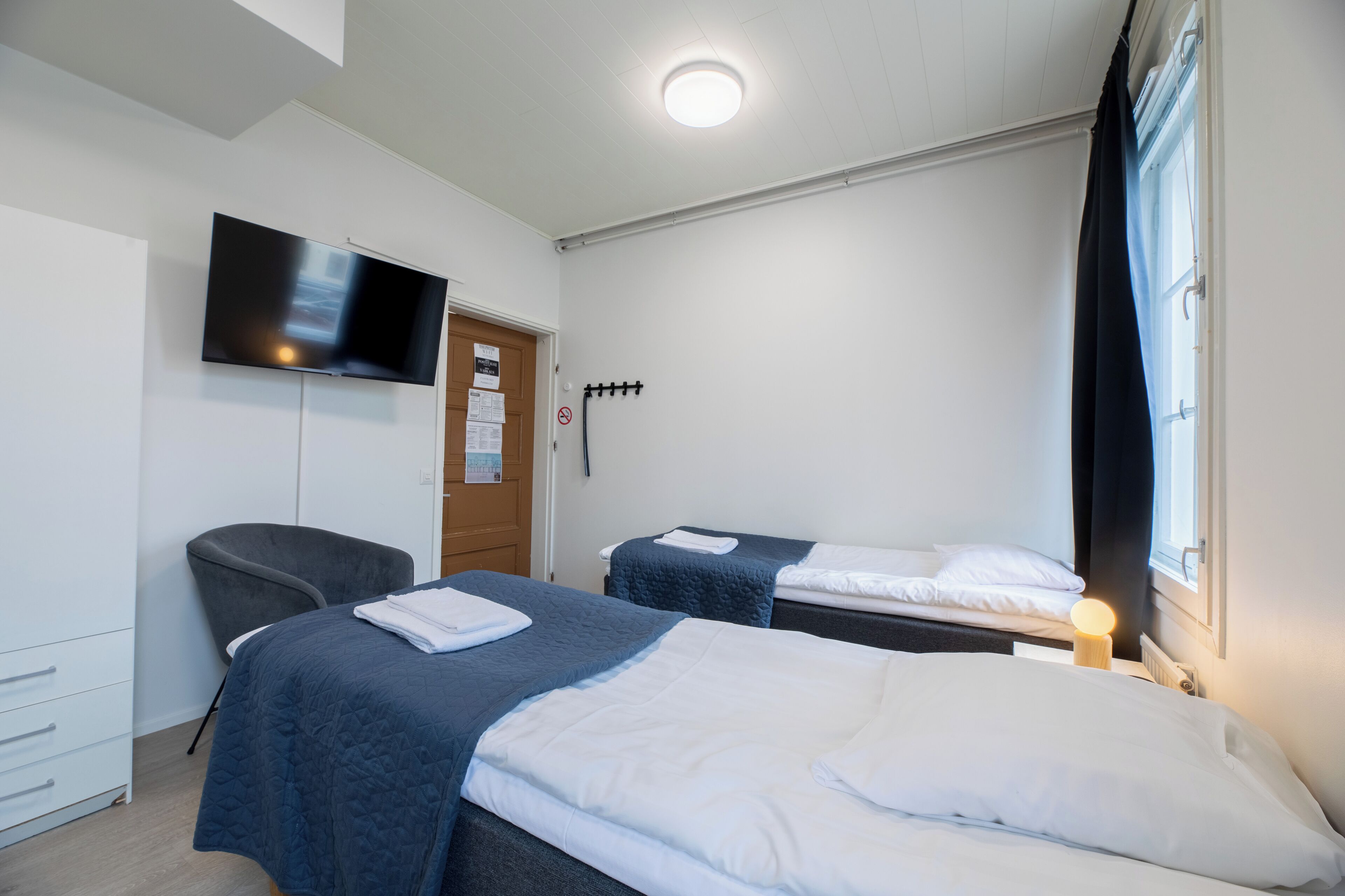Ein Hotelzimmer mit zwei Betten, einem wandmontierten Flachbildfernseher, einem Schreibtisch mit Stuhl und einem Fenster mit Vorhängen.