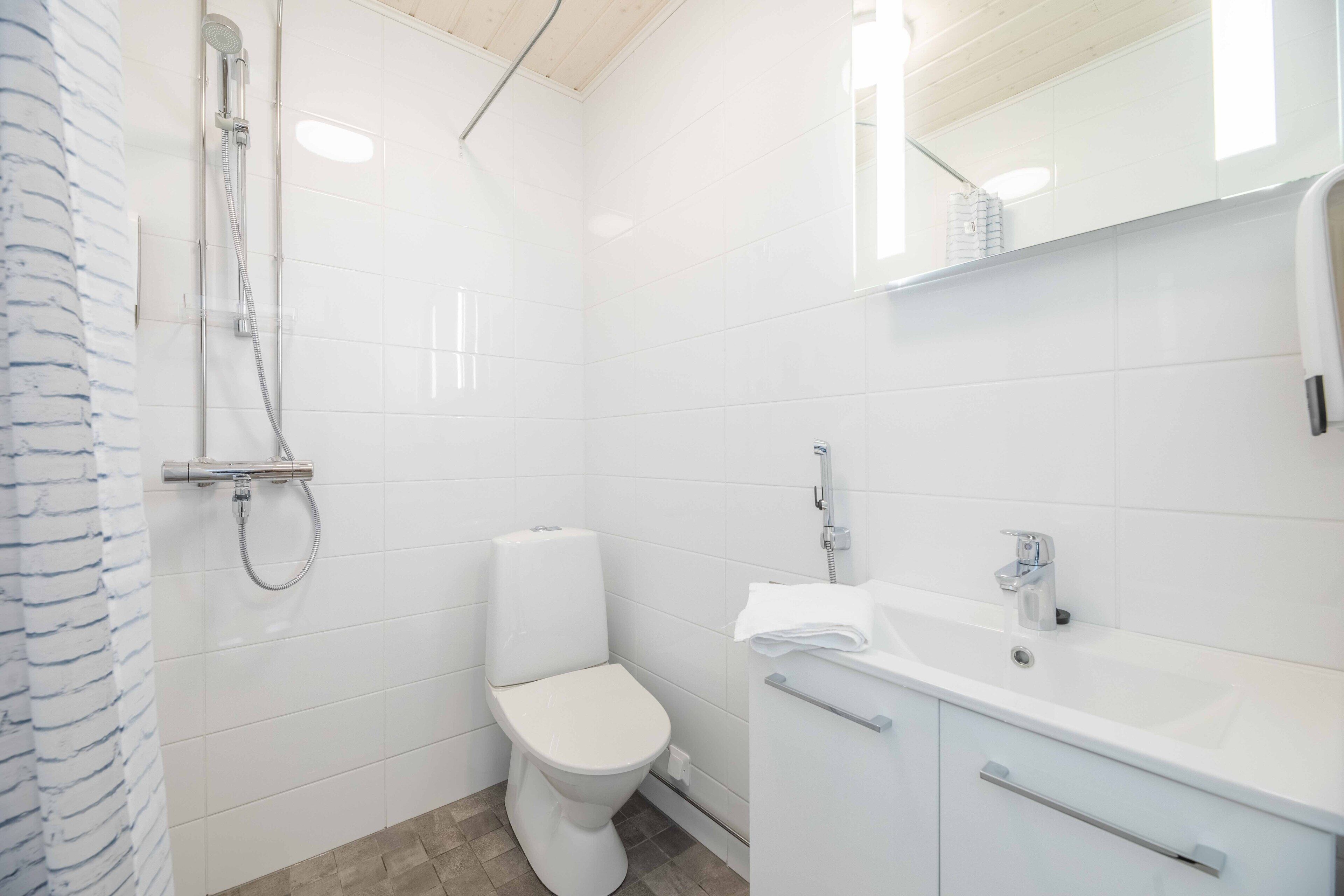A modern bathroom with white tiles, a toilet, a shower, and a sink with a mirror above it.