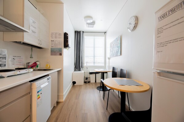 A compact kitchen with a dishwasher, a small table, and chairs. There is a window with curtains, a clock, and a refrigerator.
