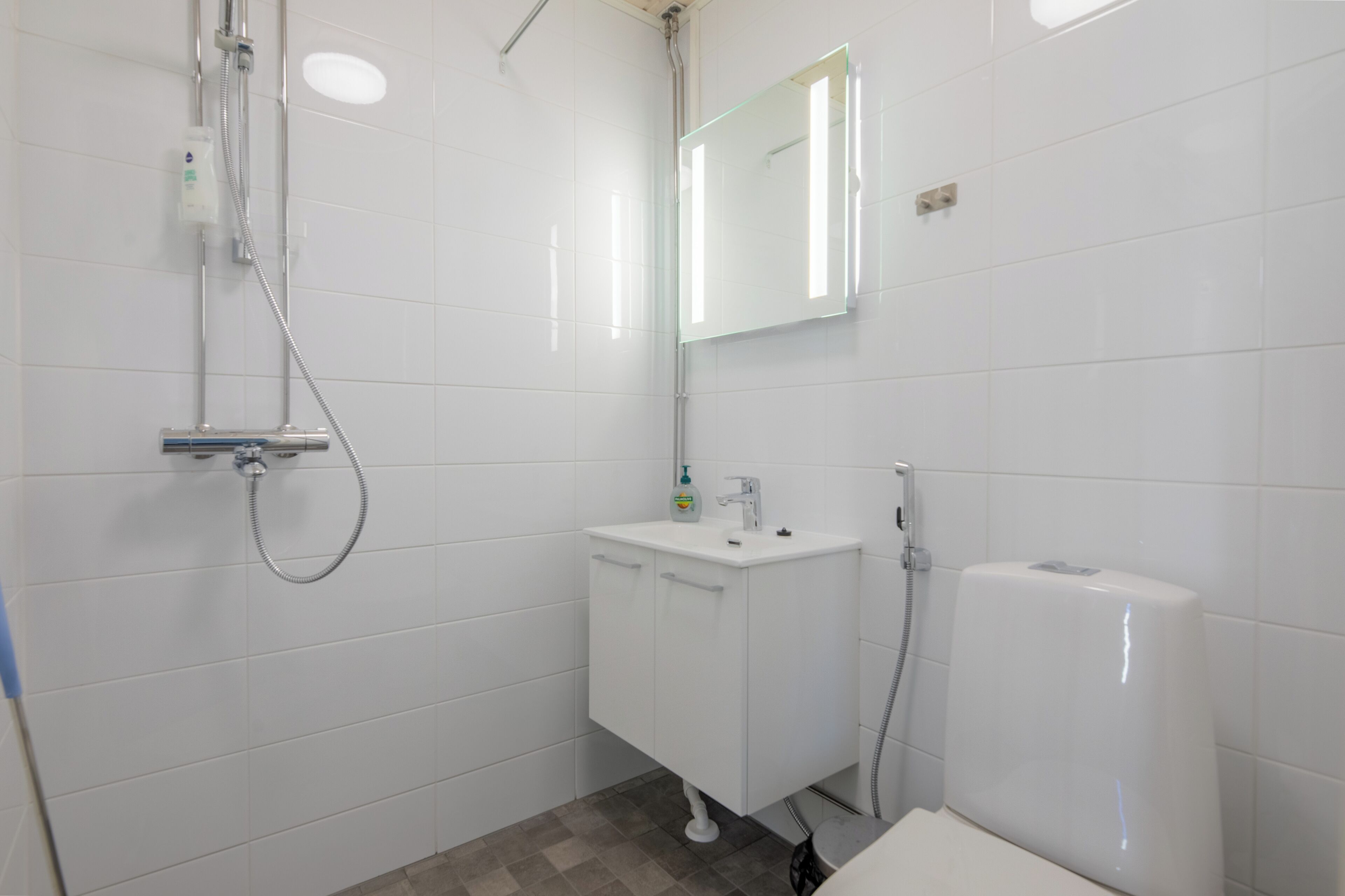 A bathroom with white tiles, a sink, a mirror, a shower, and a toilet.