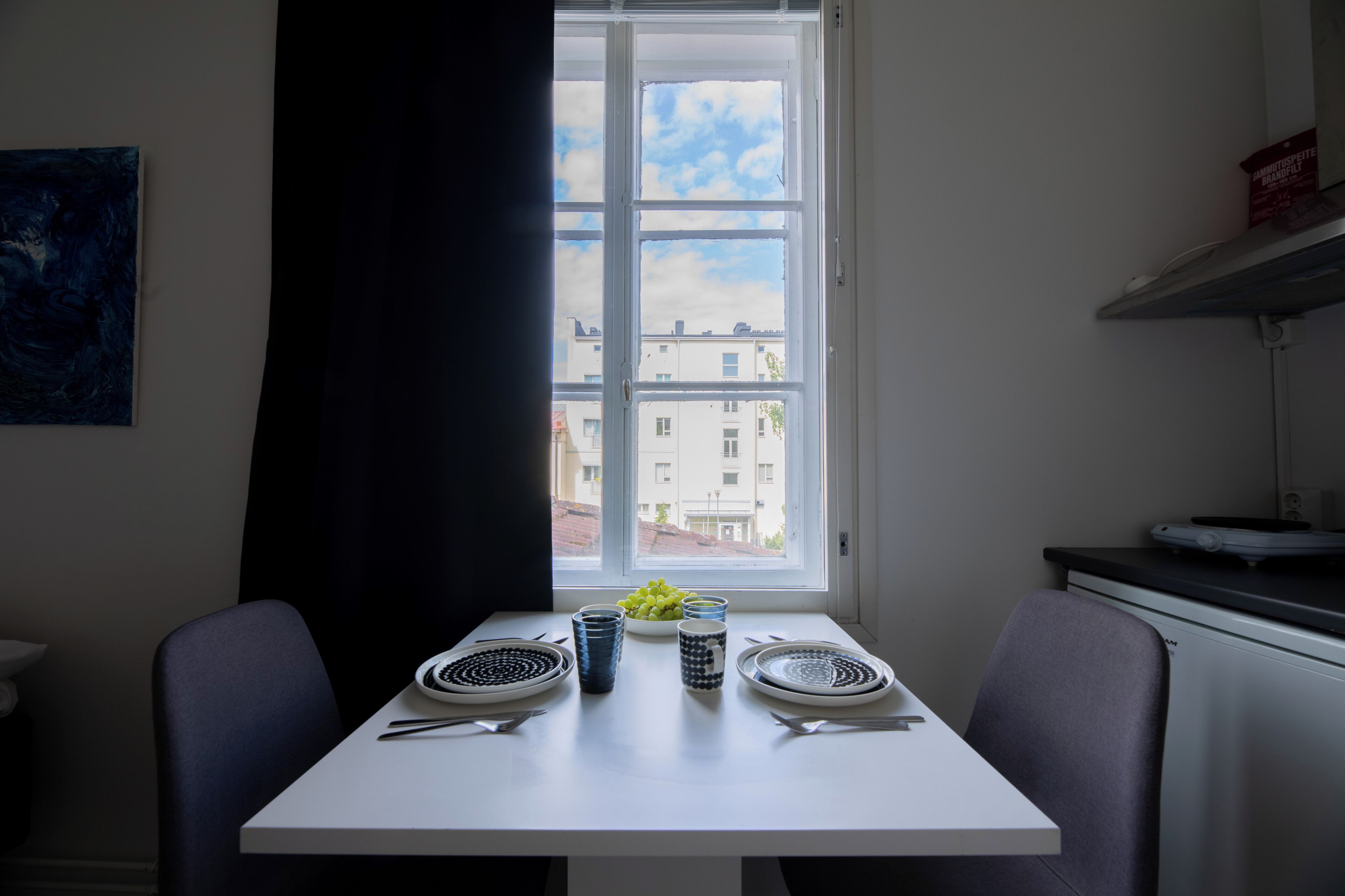 Un comedor con una mesa dispuesta para dos, una ventana con vistas a edificios y un cuadro en la pared.