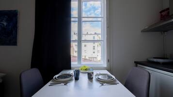 Un comedor con una mesa dispuesta para dos, una ventana con vistas a edificios y un cuadro en la pared.