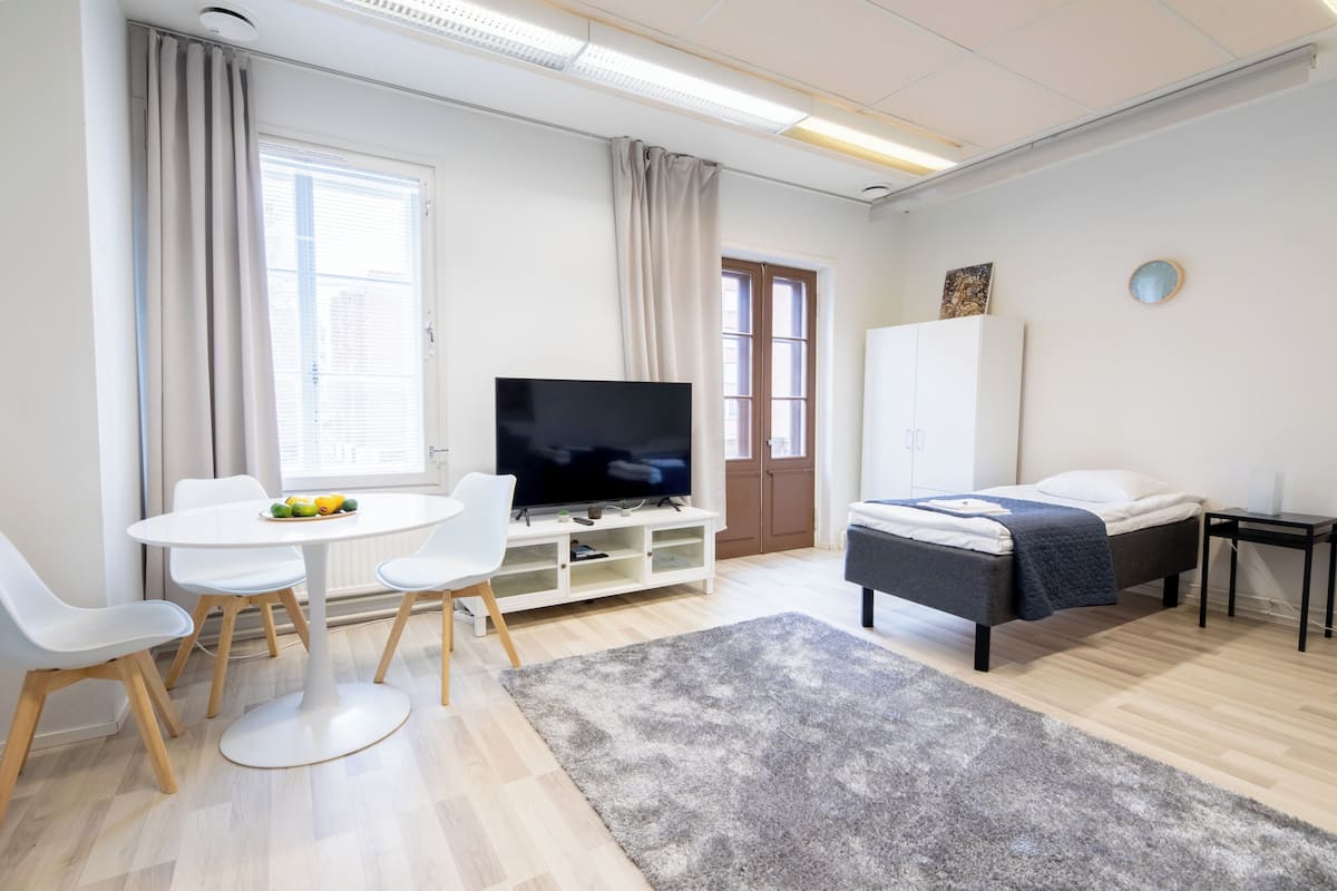 Una habitación de hotel moderna con una cama, un televisor, una mesa de comedor con sillas y un amplio ventanal con cortinas.