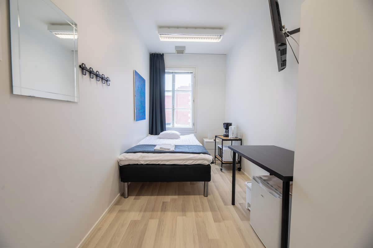 Una habitación de hotel compacta con cama, escritorio, una mesita pequeña, espejo, ventana con cortinas y aire acondicionado de pared.