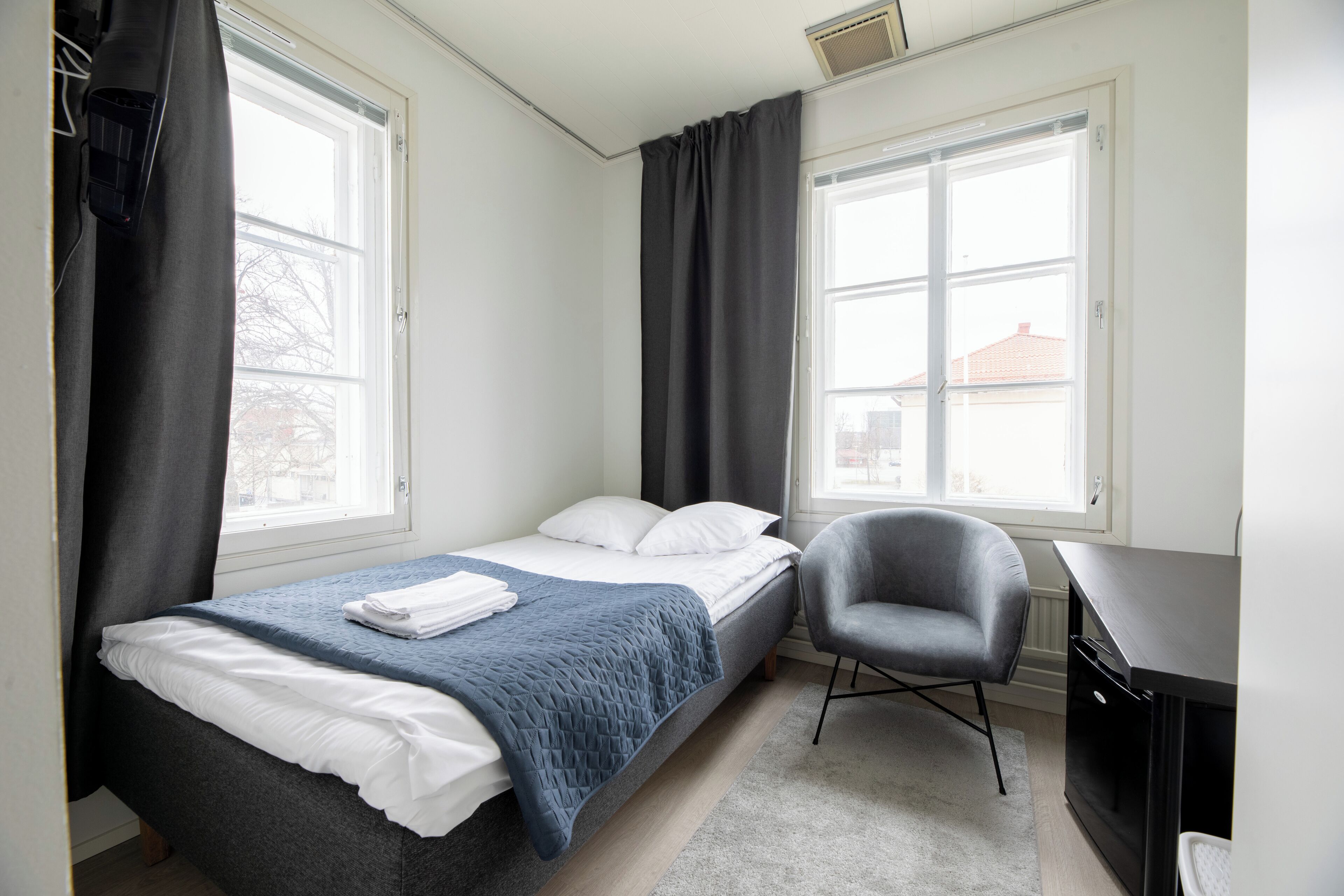 Un dormitorio con una cama, una silla, un escritorio y dos ventanas.