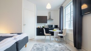 A compact hotel room with a bed, a small kitchenette, a dining area with a table and chairs, and a large window providing natural light. - Vanha Postitalo (Varkaus)