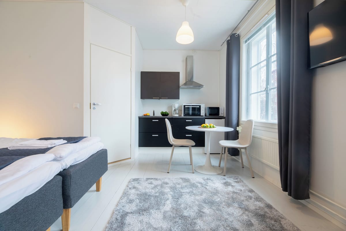 Habitación de hotel compacta con cama, kitchenette pequeña, zona de comedor con mesa y sillas, y un ventanal que proporciona luz natural.