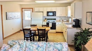 Cocina pequeña con armarios blancos, microondas, estufa y lavamanos. Cuenta con una zona de comedor con mesa y sillas.