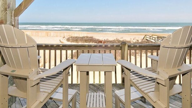 Una terraza de madera con dos sillas Adirondack y una mesita, con vista a una playa y un paseo marítimo de madera.