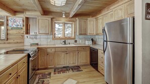 A kitchen with wooden cabinets, a stainless steel refrigerator, and a window with a view of trees. - Cook Family Lodge (Breckenridge)