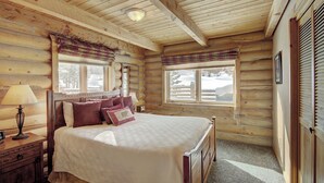 A log cabin bedroom with a bed, bedside table, lamp, and windows with curtains. - Cook Family Lodge (Breckenridge)