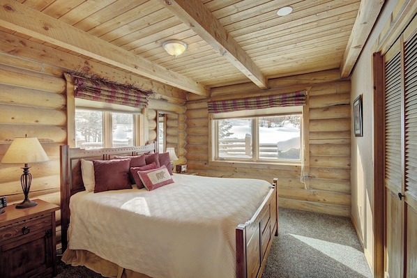 Ein Schlafzimmer in einer Blockhütte mit Bett, Nachttisch, Lampe und Fenstern mit Vorhängen.
