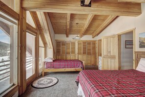 A room with wooden beams, two beds with plaid bedding, a round rug, and a view of a snowy landscape through large windows. - Cook Family Lodge (Breckenridge)