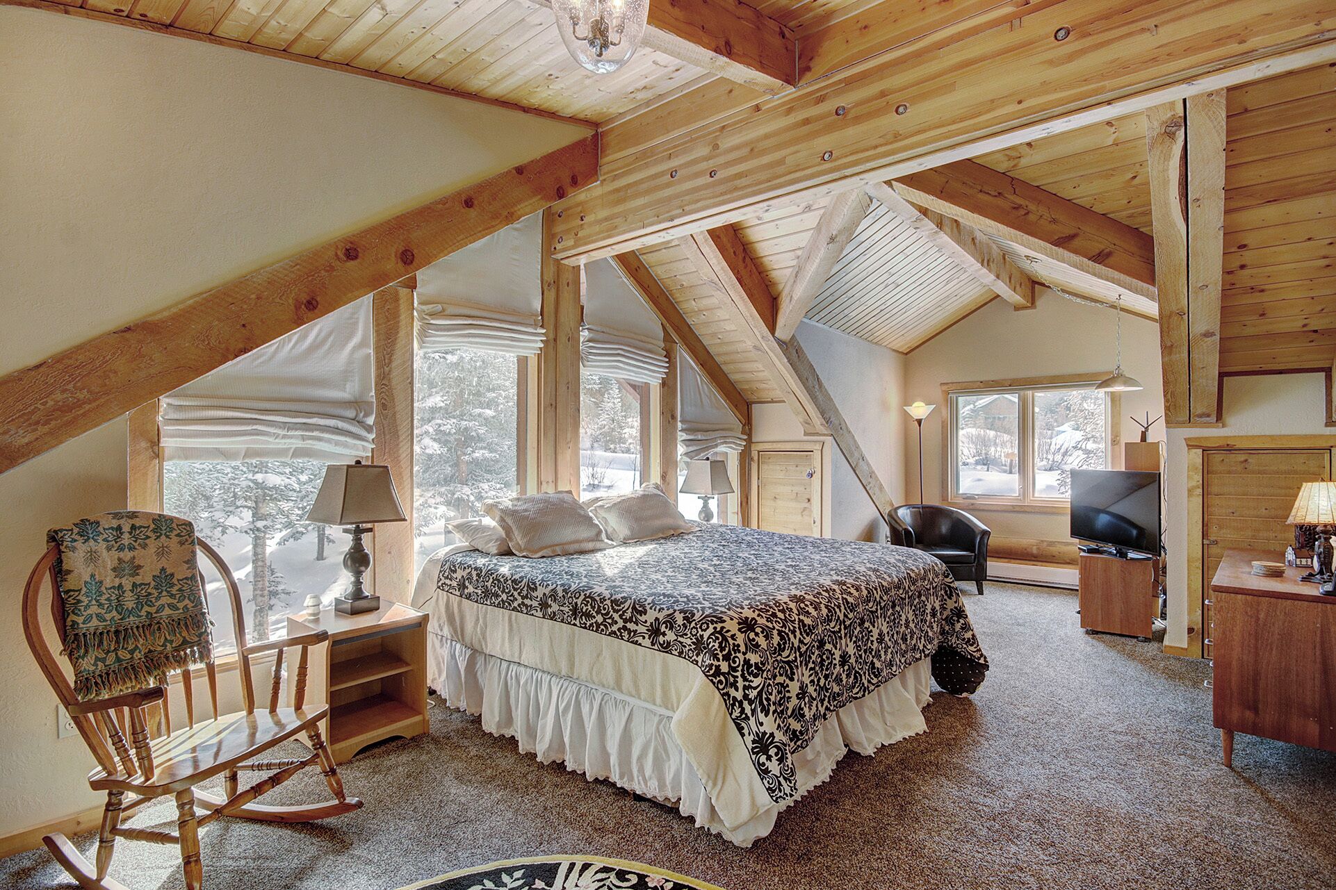 A cozy attic bedroom with a large bed, a rocking chair, a desk, and a television.