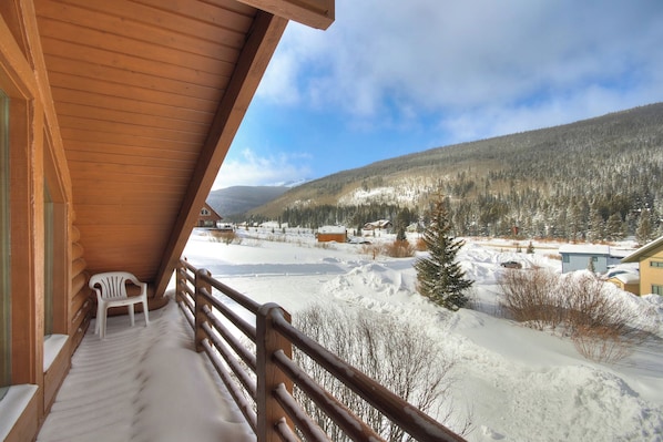 A wooden balcony with a white plastic chair, overlooking a snowy landscape with houses and a forested mountain.