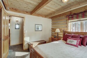 A wooden cabin bedroom with a bed, a nightstand, a wall clock, and a window with a view of trees. - Cook Family Lodge (Breckenridge)