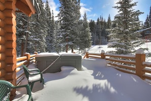 A snow-covered wooden deck with a hot tub, surrounded by snow-laden trees and a wooden fence.