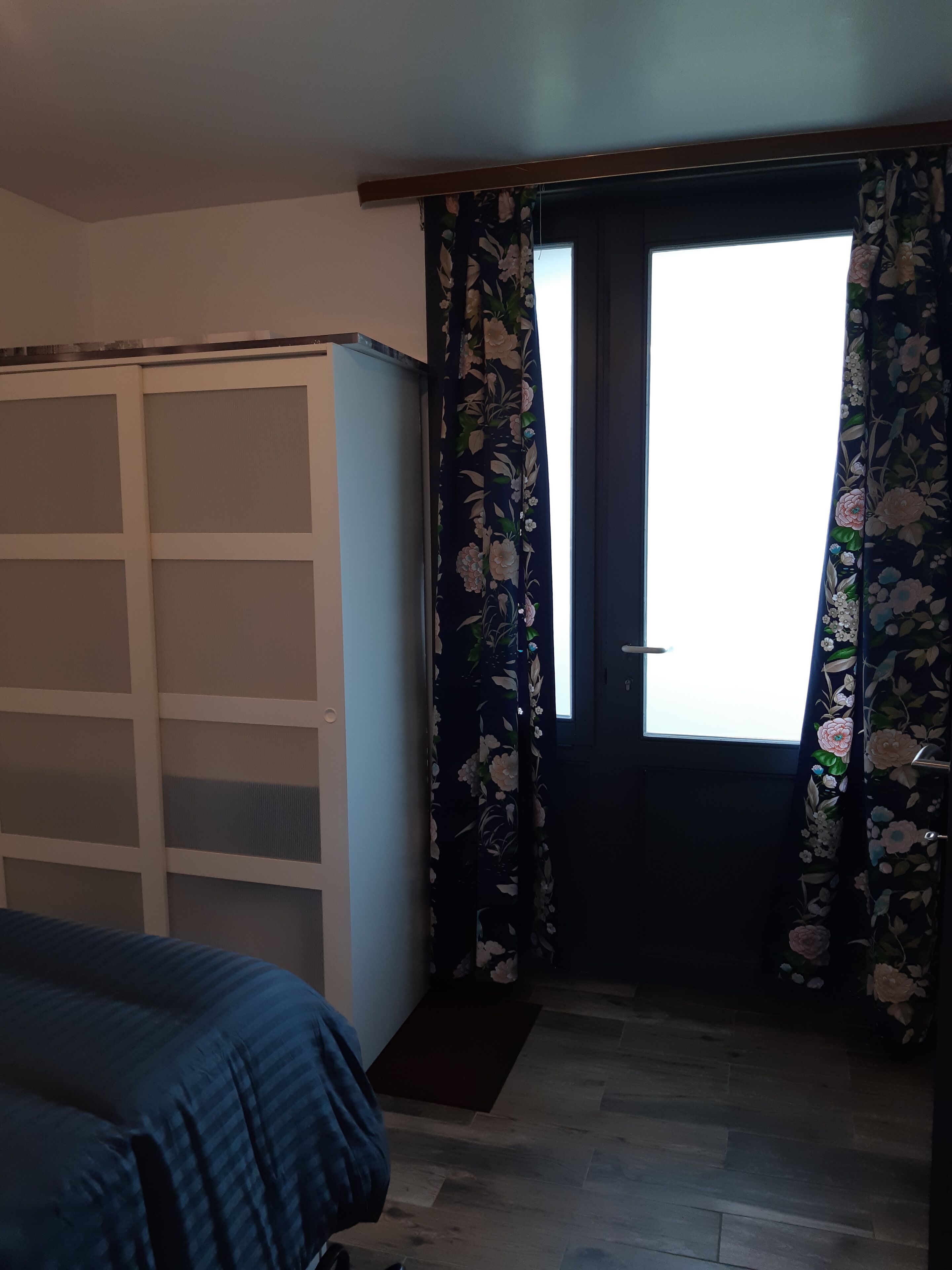 Huone, jossa on sänky, vaatekaappi, kukka-kuvioidut verhot ja ikkunasta avautuva näkymä ulos.