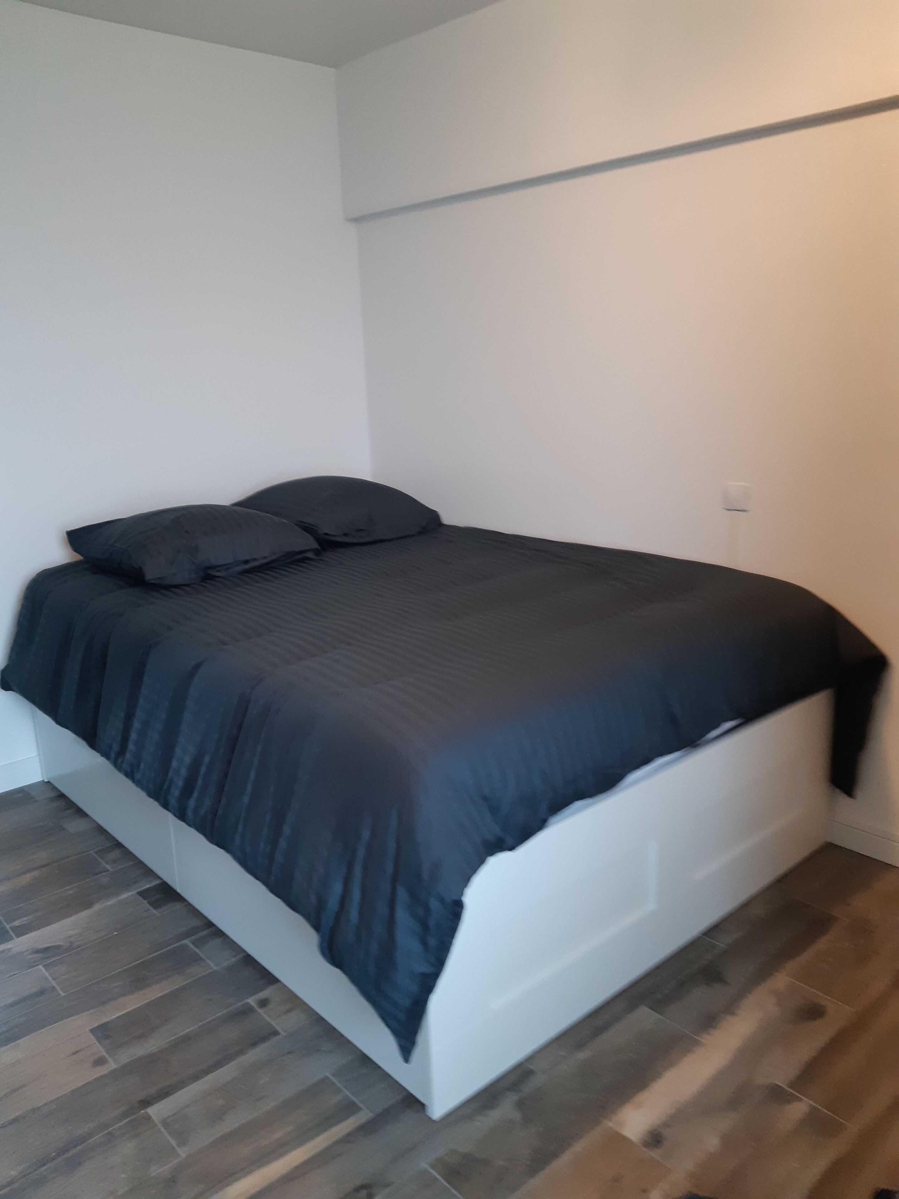 Een kleine kamer met een enkel bed, een wit hoofdbord, donkere beddengoed en houten vloeren.
