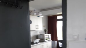 Un pasillo estrecho que conduce a una cocina con armarios blancos, un microondas y un ventanal con cortinas.