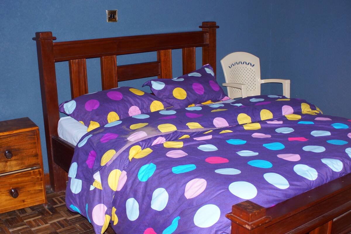 A bed with a purple polka dot bedspread, a wooden headboard, and a white chair with a perforated backrest.
