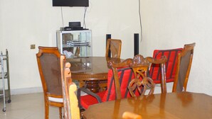 A dining area with a round wooden table, six wooden chairs with red cushions, a television mounted on the wall, and a glass cabinet with a metal shelf. - Bells Guest House (Ol Kalou)