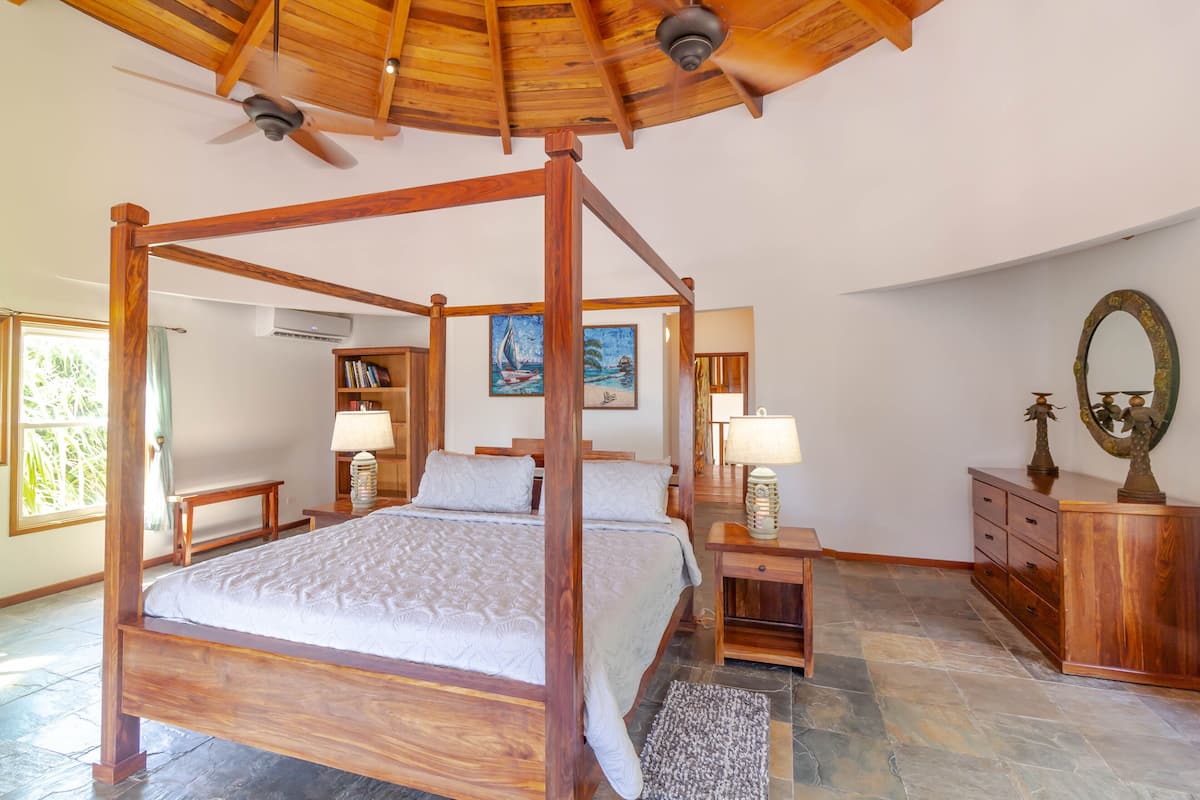 A four-poster bed with wooden headboard and footboard, flanked by nightstands with lamps, a wooden dresser, and a painting of a sailboat on the wall.