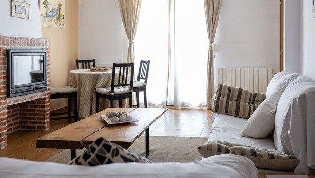 Ein gemütliches Wohnzimmer mit Kamin, einem Esstisch mit Stühlen, einer Couch und einem großen Fenster mit Vorhängen.