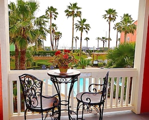 A balcony with a table and two chairs, overlooking a pool and palm trees.