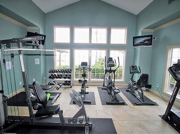 Et fitnesscenter med forskellig træningsudstyr, store vinduer og et fjernsyn.