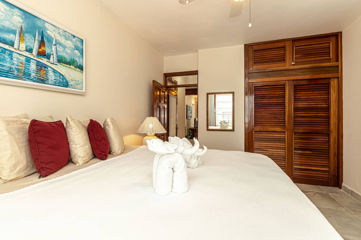 A neatly made bed with red and beige pillows, a painting of sailboats on the wall, and wooden shutters on the window.