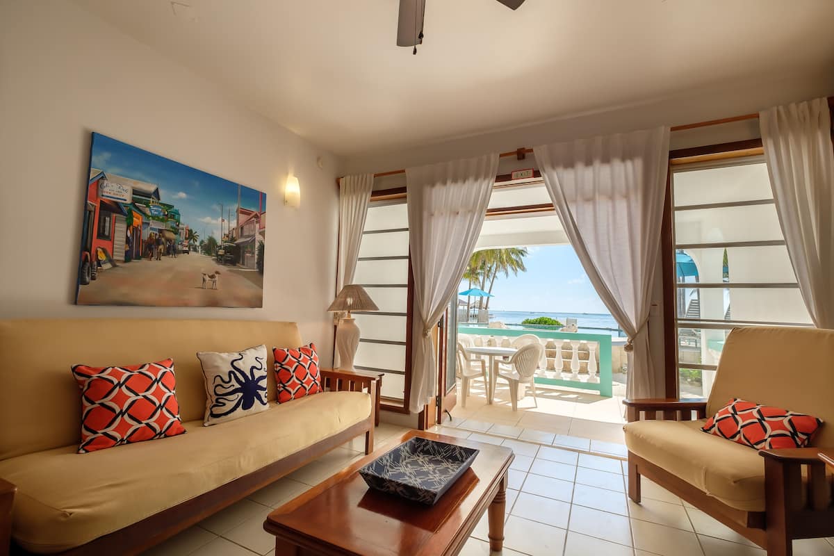 A living room with a sofa, armchairs, a coffee table, and a painting of a street scene. There is a view of a pool and palm trees through the windows.