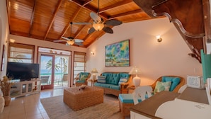 A spacious living room with a high ceiling, wooden beams, a large flat-screen TV, wicker furniture, and a view of the ocean through large windows. - Coral Bay Villas (San Pedro)