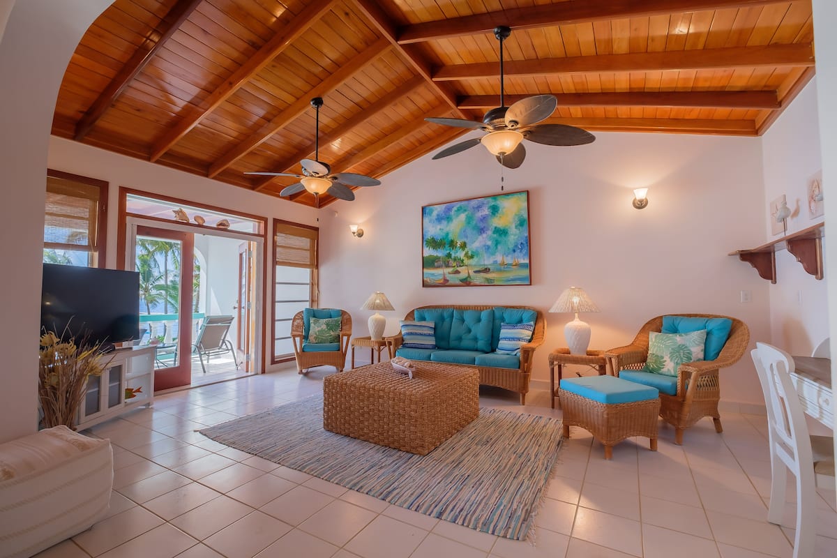 A spacious living room with a high ceiling, wooden beams, a large painting, wicker furniture, and a flat-screen TV.