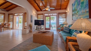A spacious living room with a wooden ceiling, a large TV, wicker furniture, and a painting of a tropical scene. - Coral Bay Villas (San Pedro)