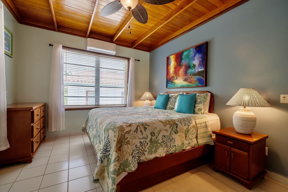 A bedroom with a bed, a nightstand, a wooden dresser, a ceiling fan, a window with blinds, and a painting on the wall.