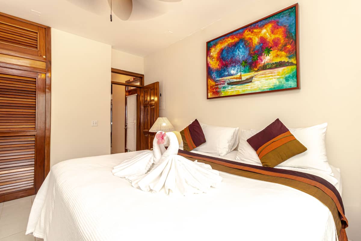 A hotel room with a neatly made bed, a colorful painting on the wall, and a wooden door.