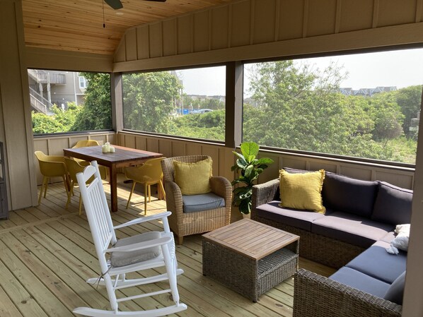 Un espacio al aire libre cubierto con mobiliario de mimbre, una mesa de madera y amplias ventanas que ofrecen vistas a la vegetación y edificios.