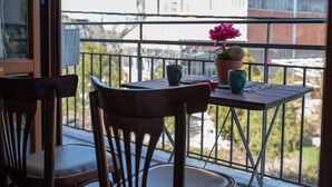 En balkon med et bord, stole, en potteplante og et vindue med udsigt over bybilledet.