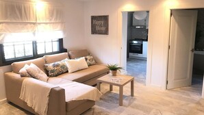Een woonkamer met een beige bank, een salontafel en een keuken die zichtbaar is door een open deur.