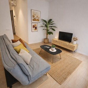 Una sala de estar con un sofá gris, una mesa de centro negra, un televisor sobre un mueble de madera y una planta grande.