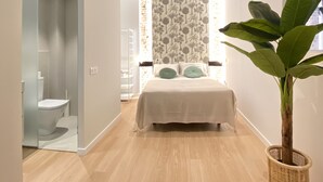 Une chambre à coucher moderne avec un grand lit, une plante en pot et une salle de bain visible à travers une porte ouverte.